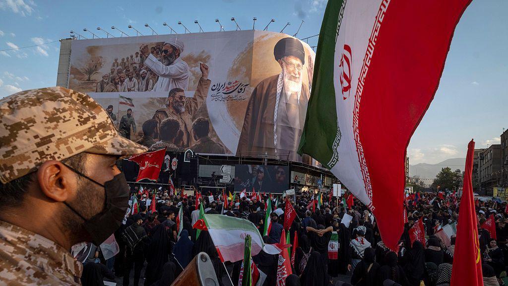 An Iranian security officer amid crowds in front of a billboard showing the late supreme leader